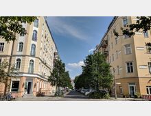 Straßenszene mit hohen, hellgelben Wohnhäusern im Altbaustil auf beiden Seiten, gesäumt von Bäumen. Im Vordergrund sind geparkte Fahrräder, ein Café mit Sitzgelegenheiten und ein schmaler Gehweg zu sehen.