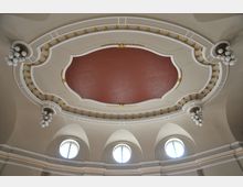 Oval geformte, reich verzierte Decke in einem historischen Gebäude, mit einem rötlichen Mittelteil, goldenen Verzierungen und weißen Ornamenten. Darunter befinden sich mehrere Rundbogenfenster, die Tageslicht einlassen.