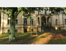 Eine historische Villa mit beigefarbener Fassade und Rundbogenfenstern, die von Grünfläche und Bäumen umgeben ist. Im Vordergrund hängt eine Schaukel an einem Baum, und eine Treppe führt zum Eingang mit Säulen und einem Balkon darüber.