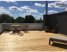 A rooftop terrace with a wooden deck featuring a table and four chairs, potted plants along one wall, and a lounge chair. The area is open to a view of trees under a partly cloudy sky.