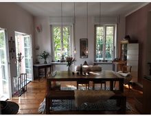 Ein heller Essbereich in einem Wohnhaus mit Holzdielenboden, großen Fenstern und einem massiven Holztisch mit Bänken, die mit Fellauflagen dekoriert sind. Der Raum ist mit einer Mischung aus modernen und antiken Möbeln sowie Blumenarrangements und hängenden Glühbirnen als Beleuchtung gestaltet.