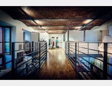 Innenansicht eines modernen Loftbüros mit langem Holzfußboden, Metallgeländern entlang eines offenen Galeriegangs und sichtbarer Ziegeldecke. Links sind große Fenster und Arbeitsbereiche zu sehen, rechts öffnet sich der Raum zu einer tieferliegenden Ebene mit Treppe und hängender Leuchte.