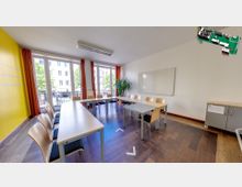 Ein moderner Seminarraum mit U-förmig angeordneten Holztischen und passenden Stühlen. Der Raum hat große Fenster mit orangefarbenen Vorhängen, eine gelbe Wand, helle Holzböden, eine weiße Tafel und eine Bürokommode.