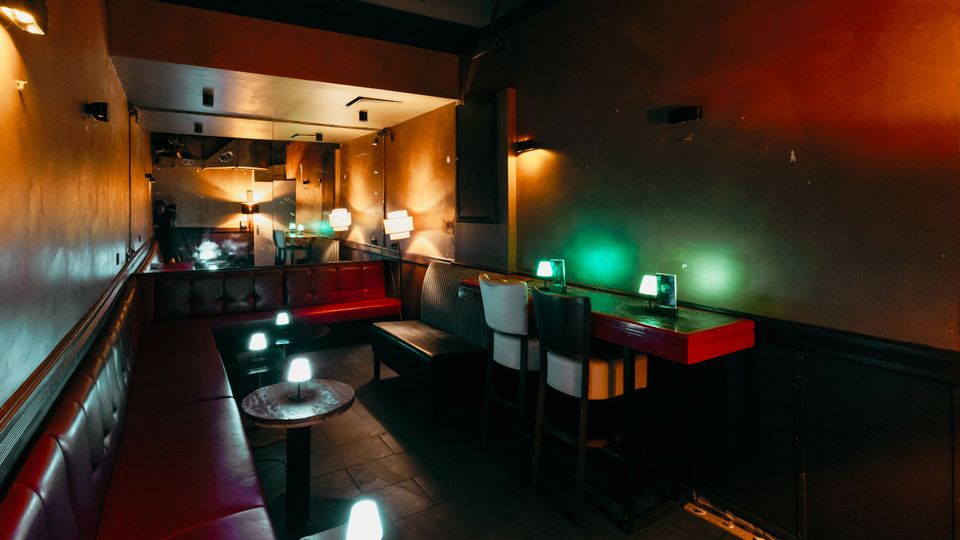 Schmaler, gedämpft beleuchteter Lounge- oder Barraum mit gepolsterten Lederbänken entlang der Wände, kleinen runden Tischen und niedrigen Tischlampen. Rechts stehen hohe Barhocker an einem Wandtisch mit farbigem Licht, im Hintergrund sind Spiegel und weitere Sitzplätze zu sehen.