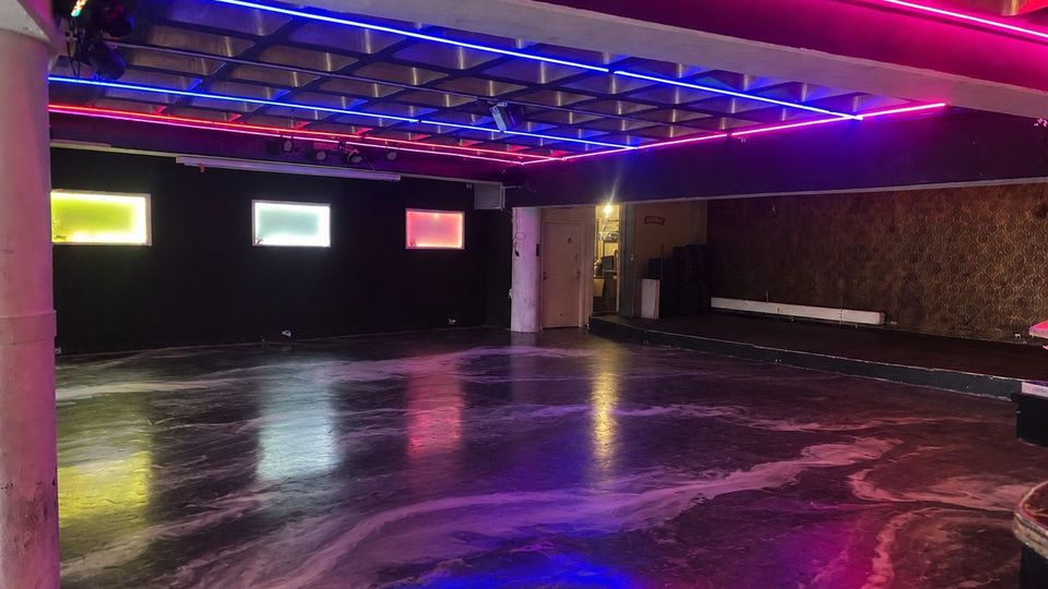 Großer, leerer Innenraum wie ein Club- oder Veranstaltungsraum mit glänzendem dunklem Boden und einer Decke mit pinken und blauen Neon-Lichtleisten. An der Rückwand befinden sich drei kleine rechteckige Fenster mit farbigem Licht, daneben eine Tür und an der rechten Wand eine lange niedrige Sitzbank vor gemusterter Tapete.