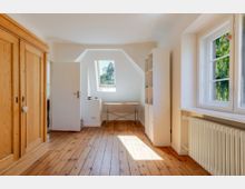 Heller Dachgeschossraum mit Holz-Dielenboden, großem Holzkleiderschrank und einem Schreibtisch unter einem Dachfenster. Rechts sorgen ein breites Seitenfenster mit Blick ins Grüne, ein Heizkörper und ein weißes Regal für eine schlichte Einrichtung.