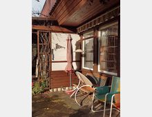 Holzterrasse mit einer Hauswand aus Holz und Putz, großer Fensterfront und Markise. Auf der Terrasse stehen ein Sonnenschirm, eine Holzbank, bunte Gartenstühle und ein Liegestuhl, umgeben von Kletterpflanzen und herbstlichem Laub.