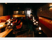 Gemütlicher Bar- oder Lounge-Innenraum mit roten gepolsterten Sitzbänken, kleinen runden Tischen und vielen niedrigen Tischlampen. Dunkle Wände und eine freiliegende Ziegelwand mit Wandleuchten prägen den Raum, dazu stehen Sessel in Reihen vor einer abgetrennten Rückwand.