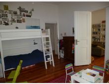 Ein Kinderzimmer mit einem weißen Hochbett, Wanddekor mit Fotos und Zeichnungen, einem kleinen Tisch und Stühlen sowie einem Holzfußboden. Rechts führt eine offene Tür in einen weiteren Raum mit Bücherregalen und einem Sofa.