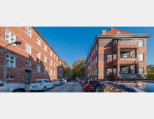 Eine enge Straße, gesäumt von mehrstöckigen roten Backsteingebäuden mit weißen Fenstern; links ist ein Wohnhaus ohne Balkone, rechts ein Gebäude mit sichtbaren Balkonen. Im Vordergrund sind geparkte Autos, im Hintergrund Bäume und ein weiteres Gebäude sichtbar.