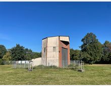 Blechverkleidetes, rostfarbenes Industriegebäude mit großem, geschlossenem Tor, umgeben von einem Metallzaun. Das Gebäude steht auf einer Wiese, im Hintergrund sind Bäume vor blauem Himmel zu sehen.