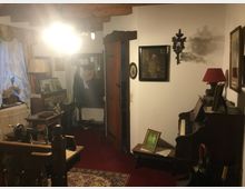 Flur in einem rustikalen Haus mit Holzbalkendecke, schummriger Beleuchtung und antiken Möbeln. Zu sehen sind eine Nähmaschine, ein Klavier, gerahmte Bilder und Dekorationsobjekte; links ein Fenster mit Spitzenvorhängen, rechts eine Holztür.