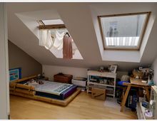 Dachgeschosszimmer mit Holzboden, zwei großen Dachfenstern und schräger Decke. Links steht ein Bambusbett mit bunter Tagesdecke, während rechts ein Schreibtisch, Regale und Umzugskartons zu sehen sind.