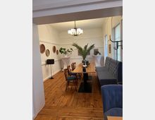 Ein gemütlicher Speisebereich mit Holzboden, einer langen Holztischreihe, gepolsterten blauen Sitzbänken und Holzstühlen. Der Raum hat weiße Wände mit dekorativen Akzenten, Pflanzen auf Säulen und moderne Wanddekorationen. Eine große, zentrale Lampe hängt von der Decke.