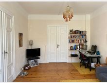 Wohnzimmer mit Holzboden, weiß gestrichenen Wänden und einer Doppeltür in klassischem Design. Links ein Fernseher auf einem kleinen weißen Regal, rechts ein Schreibtisch mit Computer, Bücherregal und Büromaterialien; darüber hängt ein dekorativer Kronleuchter.