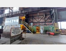 Industriehalle mit Backsteinwänden, Metallträgern und großen Fenstern; eine gelbe Treppe führt zu einer erhöhten Plattform mit Glasfenstern. Im Vordergrund ein Hinweisschild „Achtung Schrottplatz!“ und rechts eine Person in einem silbernen Schutzanzug.