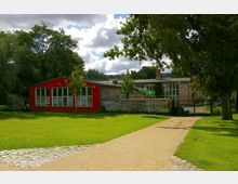 Eine grüne Parklandschaft mit einem gelblichen Kiesweg, der zu einem roten, rechteckigen Gebäude mit großen Fenstern führt. Im Hintergrund stehen weitere Gebäude, teils von Bäumen und einer niedrigen Mauer umgeben.