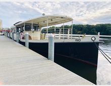 Ein kleines, weiß-schwarzes Ausflugsboot mit der Aufschrift "INO" liegt an einem hölzernen Steg, umgeben von ruhigem Wasser mit einem bewaldeten Ufer im Hintergrund unter leicht bewölktem Himmel.