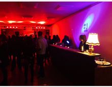 Ein Veranstaltungsraum mit gedimmtem, rotem Licht, einer Bar auf der rechten Seite und Menschen, die in Gruppen stehen. Im Hintergrund befindet sich eine Bühne mit Scheinwerfern und Musikequipment.