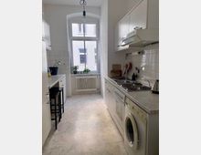 Schmale Küche in hellen Farbtönen mit weißen Schränken, einer beigen Arbeitsplatte und weißen Fliesen an der Wand. Ein großes Fenster mit Heizkörper darunter lässt Tageslicht herein, und Elektrogeräte wie Herd und Waschmaschine sind an der rechten Seite eingebaut.