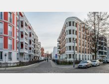 Straßenszene mit modernen, mehrstöckigen Wohngebäuden auf beiden Seiten, links mit roten und weißen Elementen, rechts mit einer Fassade aus Backstein und weißen Akzenten. Im Vordergrund sind geparkte Autos, Gehwege und einige Bäume zu sehen, während die Straße in der Mitte in die Tiefe führt.