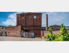 Backstein-Gebäude mit einem hohen Schornstein, umgeben von einem gepflasterten Hof; im Vordergrund befinden sich grüne Pflanzen. Das Gelände umfasst weitere niedrigere Gebäude und Mauern mit Graffiti.