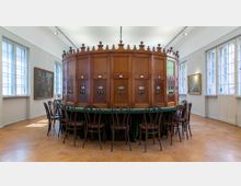 Ein historischer Raum mit einer großen, runden Holzkonstruktion in der Mitte, die mit nummerierten Sichtfenstern und Aussparungen ausgestattet ist, umgeben von einer Reihe dunkler Holzstühle an einem grünen, stoffbezogenen Tisch. Der Raum verfügt über hohe Fenster, Gemälde an den Wänden und einen Holzboden.
