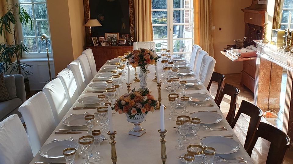 Langer Speisesaal mit großem, weiß gedecktem Banketttisch, zahlreichen Gedecken, Kristallgläsern mit Goldrand, Kerzenleuchtern und Blumenarrangements. Helle Wände, hohe Fenster und eine Glastür mit Vorhängen sowie ein Kamin und ein großes gerahmtes Porträt prägen den Raum.
