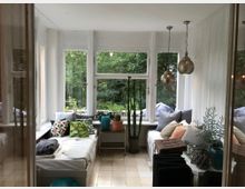 Heller Wintergarten mit großen Fenstern und Blick auf grüne Bäume, eingerichtet mit weißen Sitzflächen, bunten Kissen, Decken und Pflanzen in Töpfen. Von der Decke hängen zwei dekorative Lampen, und ein kleiner Beistelltisch steht in der Mitte.