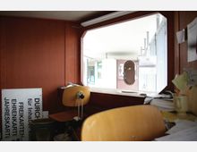 Holzvertäfelter Innenraum eines kleinen Ticketschalters mit Schreibtisch, Holzstühlen und verstreuten Papieren. Durch das große Fenster ist ein überdachter Außenbereich mit einer offenen Klappe und Gebäudeelementen zu sehen.