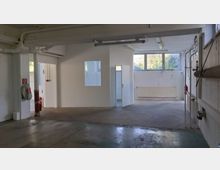 Innenraum eines leerstehenden Gebäudes mit weißen Wänden, großem Fenster, sichtbaren Rohren und einer Heizung unter dem Fenster. Auf der linken Seite befinden sich ein Feuerlöscher und Feuerschutzausrüstung, rechts verlaufen Rohre an der Wand.