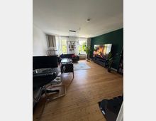 Helles Wohnzimmer mit breiten Holz-Dielen, dunkler Akzentwand und großem Flachbildfernseher auf einem Lowboard. Vor zwei großen Fenstern mit Vorhängen stehen Sofa und Couchtisch auf einem Teppich; links ist ein Arbeitsplatz mit Schreibtisch, Monitoren und Stuhl zu sehen.