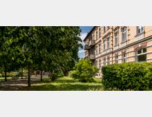 Mehrstöckiges historisches Gebäude mit gelblichem Ziegelmauerwerk und großen Fenstern, daneben ein grüner Park mit Bäumen, Büschen, Wiesen und Sitzbänken auf einem gepflasterten Weg.