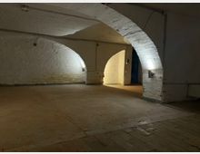 Gewölbter Kellerraum mit weiß gestrichenen Backsteinwänden und großen Rundbögen, beleuchtet durch wandmontierte Lampen. Der Raum hat einen glatten Betonboden und führt durch einen Bogen in einen helleren Nebenbereich.