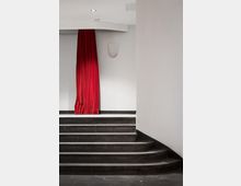 Ein schlichter Innenraum mit einer grauen Treppe, die zu einer weißen Wand mit einem roten Vorhang führt. Die Wand ist mit einer kleinen, abgerundeten Wandleuchte geschmückt.