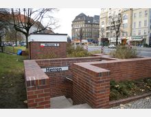 Backsteintreppe zu einer öffentlichen Herrentoilette im Außenbereich, umgeben von niedrigen Backsteinmauern und einer kleinen Grünfläche; im Hintergrund mehrstöckige Gebäude mit Geschäften an einer belebten Straße.