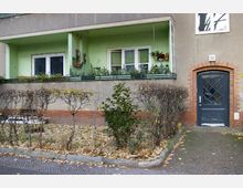 Wohnhausfassade mit grün gestrichener Balkonfläche, dekoriert mit Pflanzen. Links ein kleiner Vorgarten mit niedrigen Hecken und Herbstlaub, rechts eine schwarze Eingangstür mit Backsteinrahmung und der Hausnummer 23.