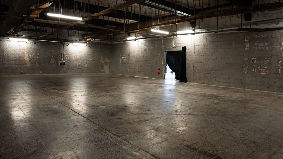 Großer, leerer Industriehallenraum mit grauen Betonblockwänden und poliertem Betonboden. Offene Decke mit sichtbaren Trägern, Kabeln und Lüftungsrohren sowie hängenden Leuchtstoffröhren; seitlich eine Türöffnung mit schwarzem Vorhang.