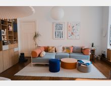 Bright modern living room with white walls, a light blue sectional sofa with colorful cushions, and round ottomans and a low coffee table on a white rug over dark wood floors. Built-in wooden shelving, a white door, potted plants, and two framed abstract artworks hang beneath a round pendant light.