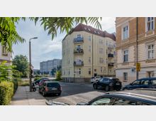 Straßenszene mit mehrstöckigen Wohngebäuden im historischen Stil, hellgelb und cremefarben gestrichen, mit Balkonen. Geparkte Autos, ein Gehweg und Bäume im Hintergrund; eine Ampel und Verkehrszeichen sind sichtbar, mit weiteren modernen Gebäuden in der Ferne.
