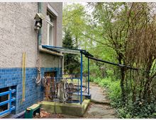 Seitenansicht eines Gebäudes mit rau verputzter Fassade und blauer Fliesenverkleidung im unteren Bereich. Im Vordergrund befinden sich Geländer mit aufgerollten Seilen, eine gelbe Rohrleitung und ein schmaler gepflasterter Weg, der in eine grüne, stark bewachsene Gartenlandschaft führt.