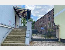 Steintreppe mit Metallgeländer führt zu einem Eingang eines hellblauen Gebäudes mit Vordach, rechts daneben ein Metalltor, das zu einem Hof mit Kopfsteinpflaster und einem Backsteingebäude führt. Im Hintergrund sind Bäume und Himmel sichtbar.