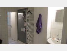 Ein schlichtes Badezimmer mit weiß gefliesten Wänden. Zu sehen sind ein Waschbecken mit einem kleinen Spiegel darüber, ein Handtuchheizkörper mit einem lila Handtuch und eine Tür, die in einen angrenzenden Raum führt.