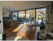 Modernes Wohnzimmer mit offener Küche, Holzboden und großer Glasfront, die auf eine Terrasse mit Palmen und Blick auf einen verschneiten Garten führt. Ein Esstisch mit weißen Stühlen und eine Kücheninsel mit Barhockern dominieren den Raum, der von Tageslicht erhellt wird.