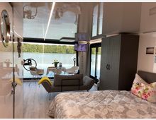 Modernes, lichtdurchflutetes Zimmer auf einem Hausboot mit Blick auf einen See und eine bewaldete Uferlandschaft. Im Raum befinden sich ein Bett mit floralem Bettbezug, eine Couch, ein kleiner Esstisch mit Stühlen, ein großer Kleiderschrank und Panoramafenster, die das Wasser und die Natur sichtbar machen.