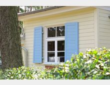 Gelbes Holzhaus mit einem weißen Fenster, das blaue Klappläden hat. Im Vordergrund befinden sich grüne Büsche und ein großer Baumstamm.