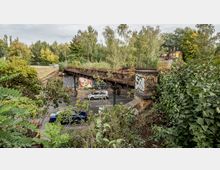 Eine alte, überwucherte Eisenbahnbrücke aus Metall und Backsteinsäulen überquert eine Straße mit fahrenden Autos. Die Umgebung ist von dichtem Grün und Bäumen umgeben, während Graffiti und Werbetafeln an den Brückenpfeilern sichtbar sind.