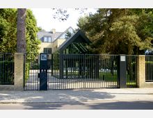 Moderne Wohnhausanlage hinter einem schwarzen Metalltor, umgeben von Bäumen und Ziegelmauern. Das Gebäude hat eine Glasüberdachung an der Vorderseite und besteht aus gelblichen Ziegeln mit großen Fenstern.