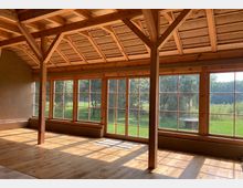 Innenansicht eines Raumes mit Holzrahmenkonstruktion, großen bodentiefen Fenstern und hölzernem Boden. Durch die Fenster ist eine grüne Wiese mit Bäumen sichtbar, die von Sonnenlicht beleuchtet wird.