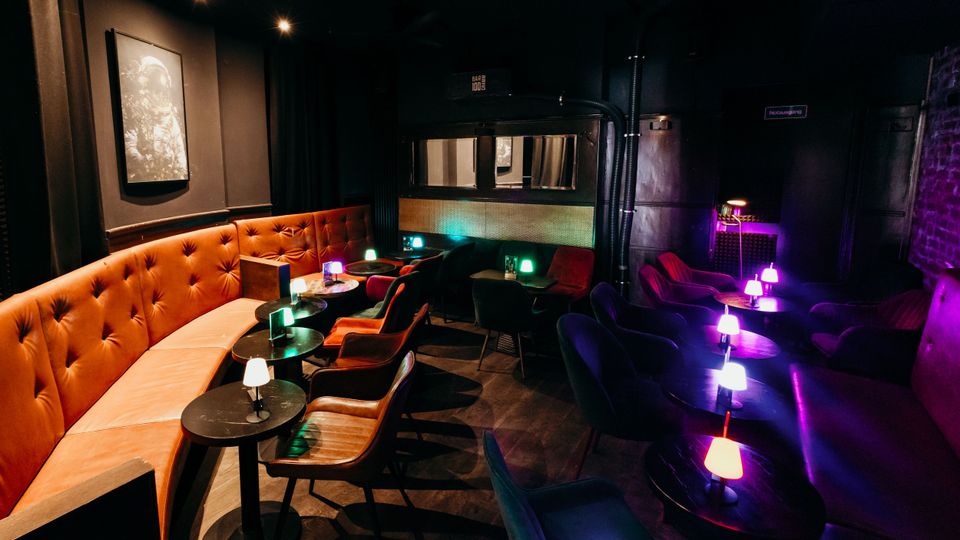 Innenraum einer dunklen Bar-Lounge mit gepolsterter, orangefarbener Sitzbank entlang der Wand, kleinen runden Tischen und Sesseln. Farbige Tischlampen und gedämpfte Deckenbeleuchtung setzen Akzente, im Hintergrund sind eine abgetrennte Nische und ein Notausgangsschild zu sehen.